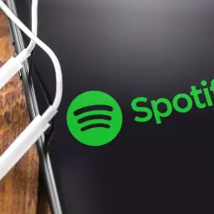 Logo Spotify na ekranie telefonu z białymi słuchawkami na drewnianym tle.
